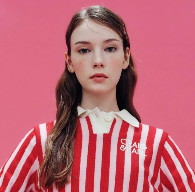 Close-up of a fair-skinned young woman with freckles, wearing a red and white striped shirt with a white collar against a pink backdrop.