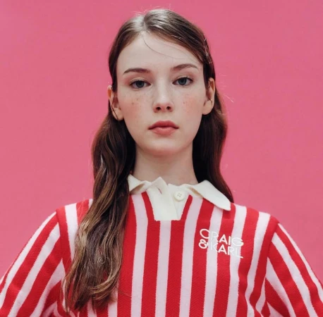 Close-up of a fair-skinned young woman with freckles, wearing a red and white striped shirt with a white collar against a pink backdrop.