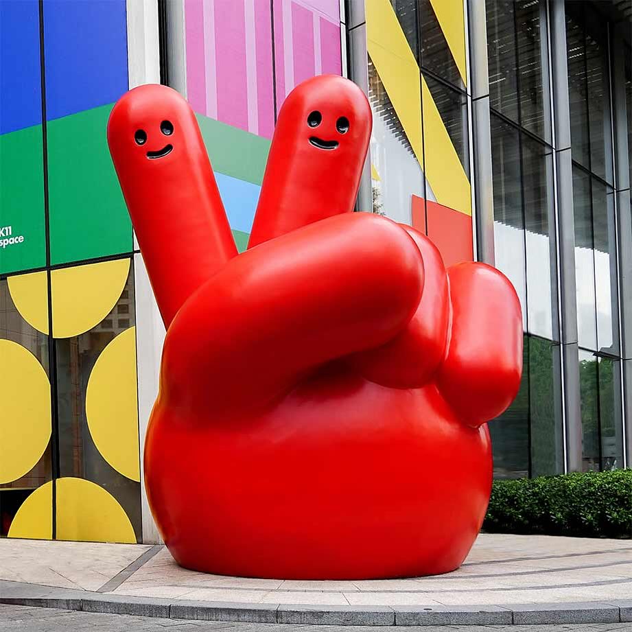 Oversized red hand sculpture with a peace sign against a building featuring colorful square and glass window design elements.