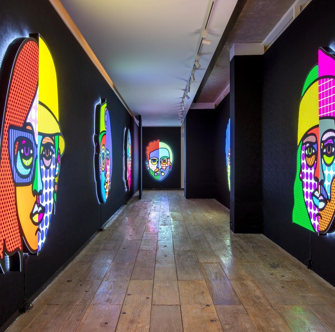 Hallway art installation with illuminated, colorful pop-art style face illustrations on black walls, and a wood-planked floor.