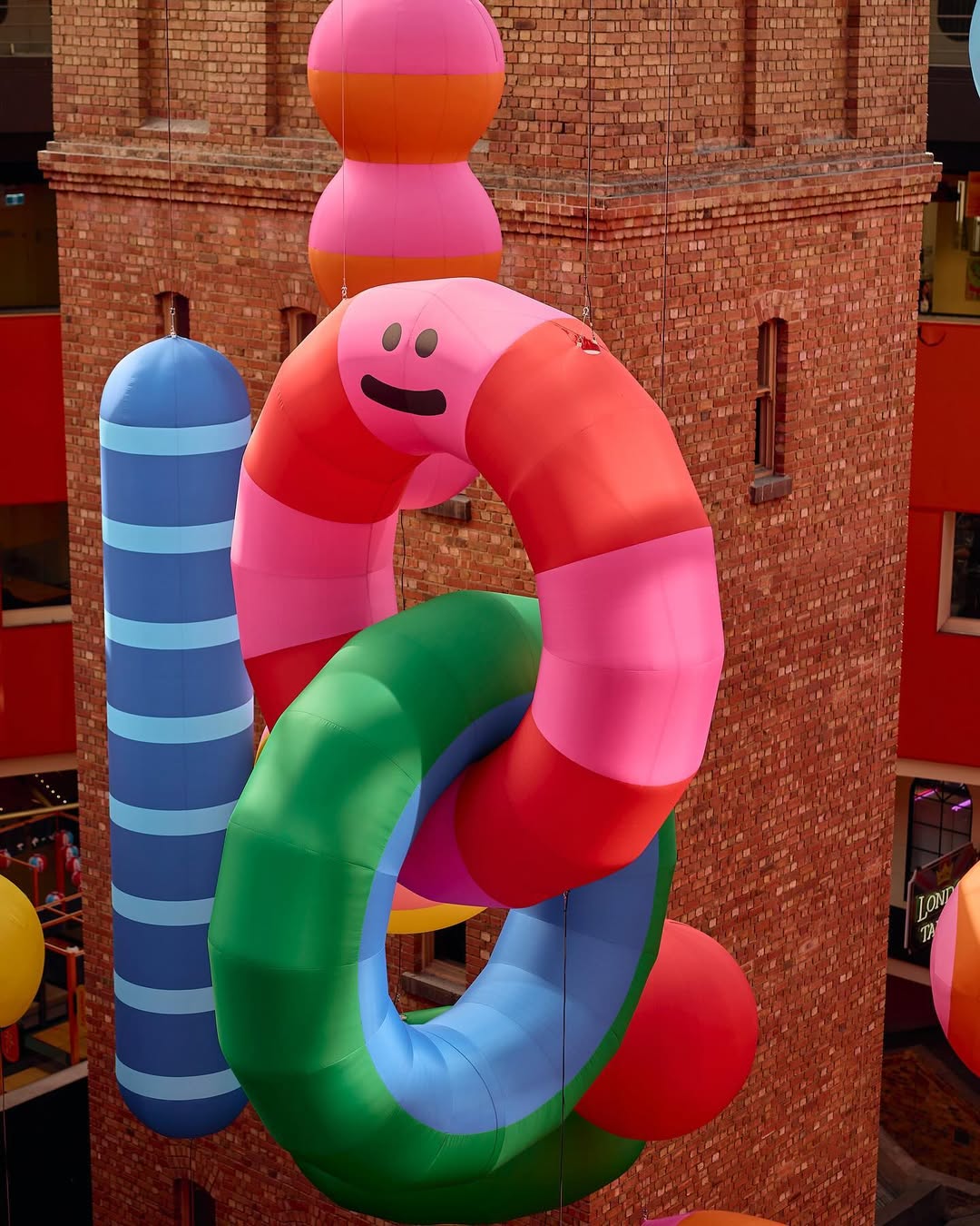 Colorful inflatable art installation featuring abstract shapes in pink, green, blue, and red hanging outside a brick building in daylight.