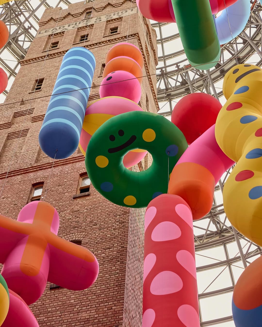 Eye-level view captures a tall brick building behind colorful inflatable objects: donuts, spheres, cylinders, and plus sign shape.