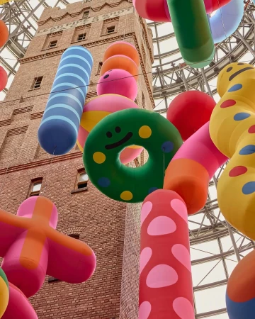 Eye-level view captures a tall brick building behind colorful inflatable objects: donuts, spheres, cylinders, and plus sign shape.