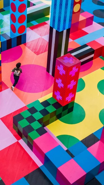 A single person walks through a colorful geometric art installation with cube and column shapes in pink, green, yellow, red, and blue.