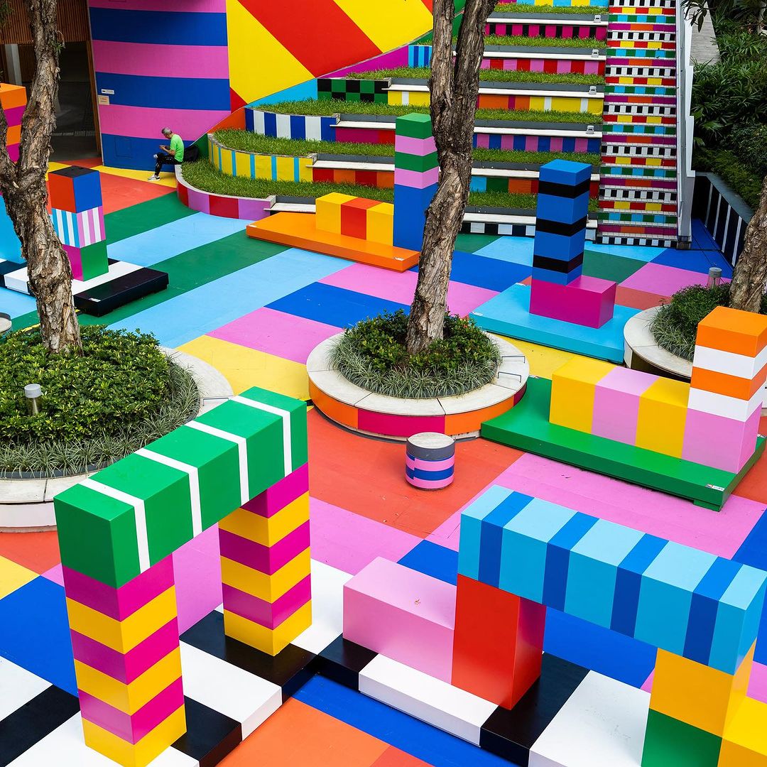 Colorful outdoor art installation with geometric shapes and trees, featuring vibrant colors like pink, blue, yellow, and green on the ground.