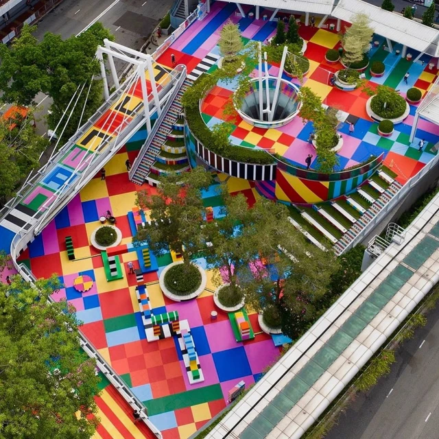 Aerial view of a colorful urban park with a pedestrian bridge, featuring a checkerboard ground and circular central structure.