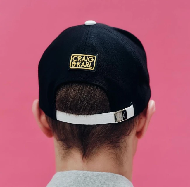 Back view of a person wearing a black and white baseball cap with a gold and black Craig & Karl logo patch against a pink background.