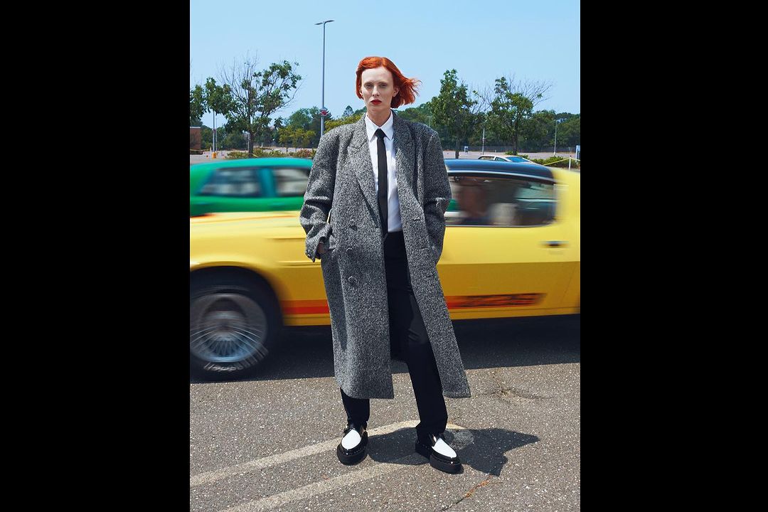 A red-haired woman in a grey coat stands in a parking lot with a blurred yellow car passing, set against a blue sky backdrop.