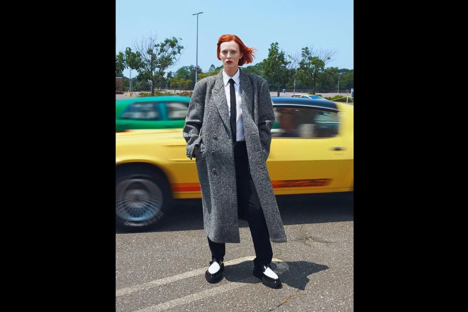 A red-haired woman in a grey coat stands in a parking lot with a blurred yellow car passing, set against a blue sky backdrop.