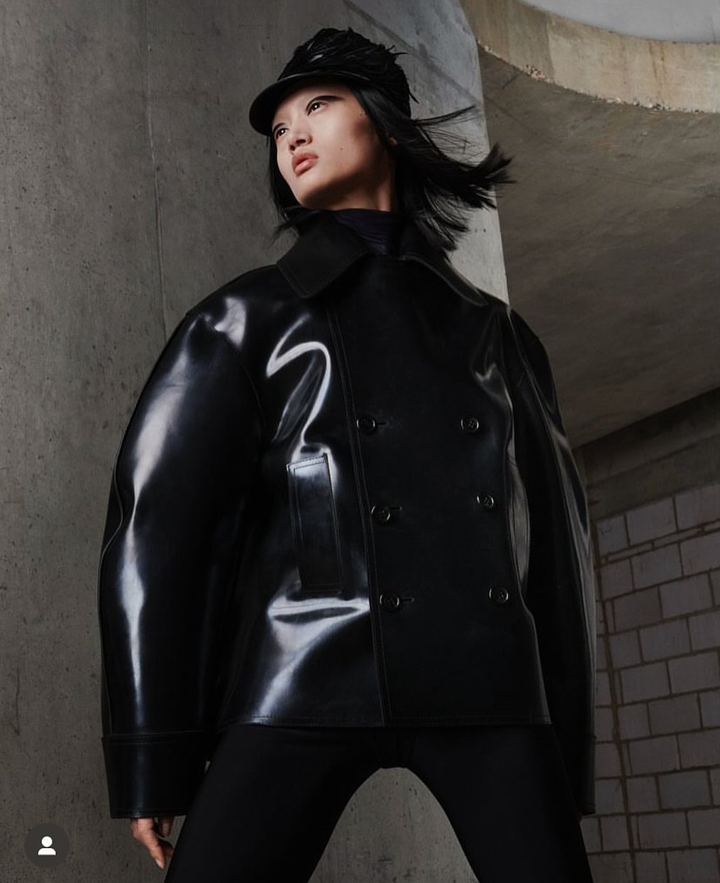 A stylish woman in a black vinyl jacket and cap stands against a textured concrete wall, showcasing modern fashion and urban aesthetics.
