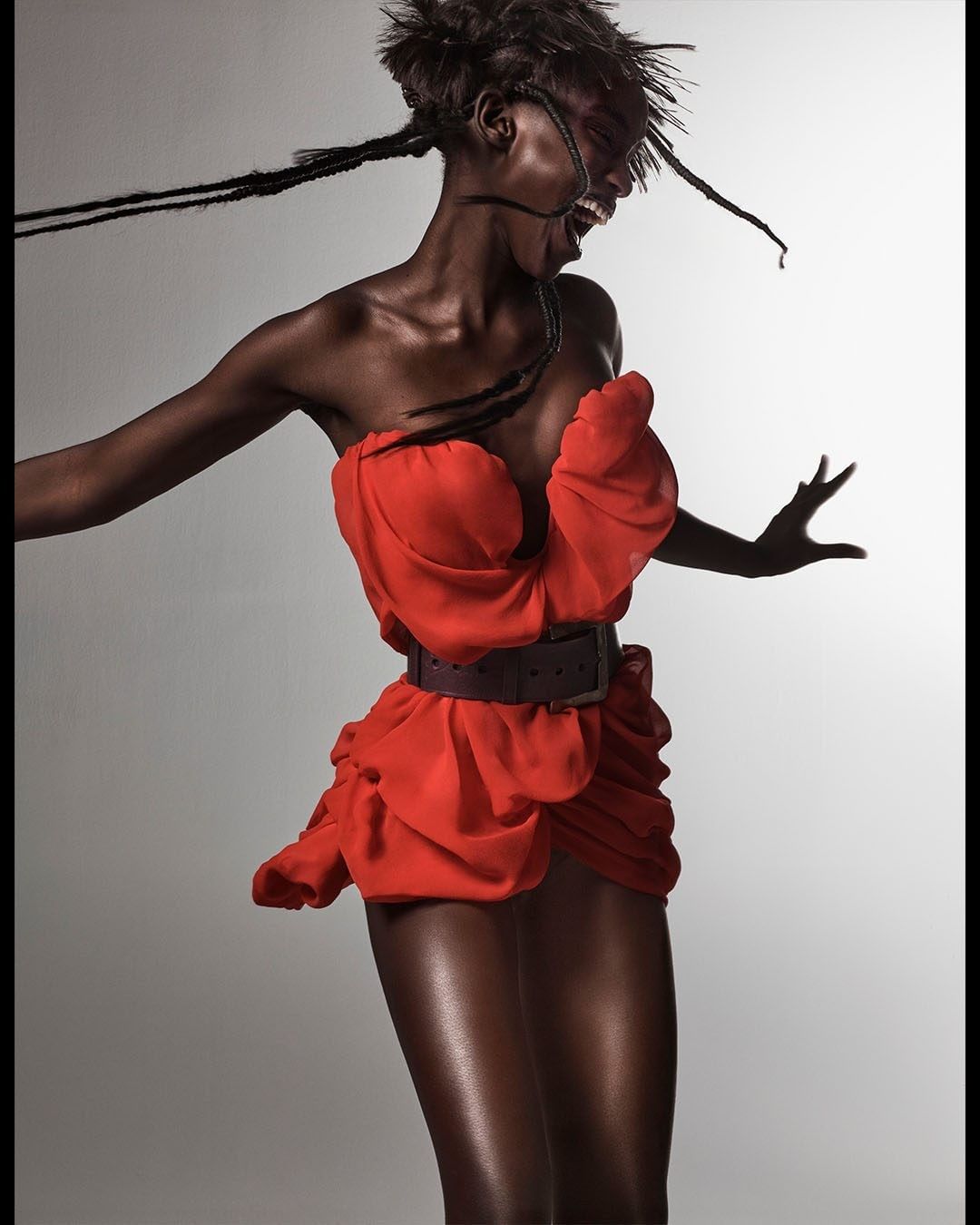 A slender African American woman in an orange dress and dark belt is captured in a studio pose against a light gray backdrop.