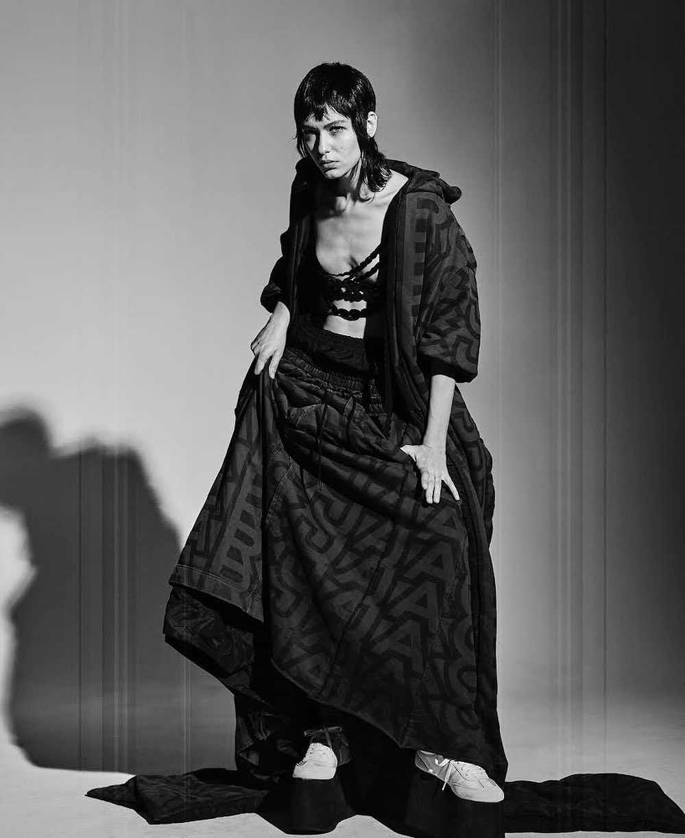 A single woman posing in a black bra, skirt, and jacket with textured patterns. The photo is a black and white studio shot.