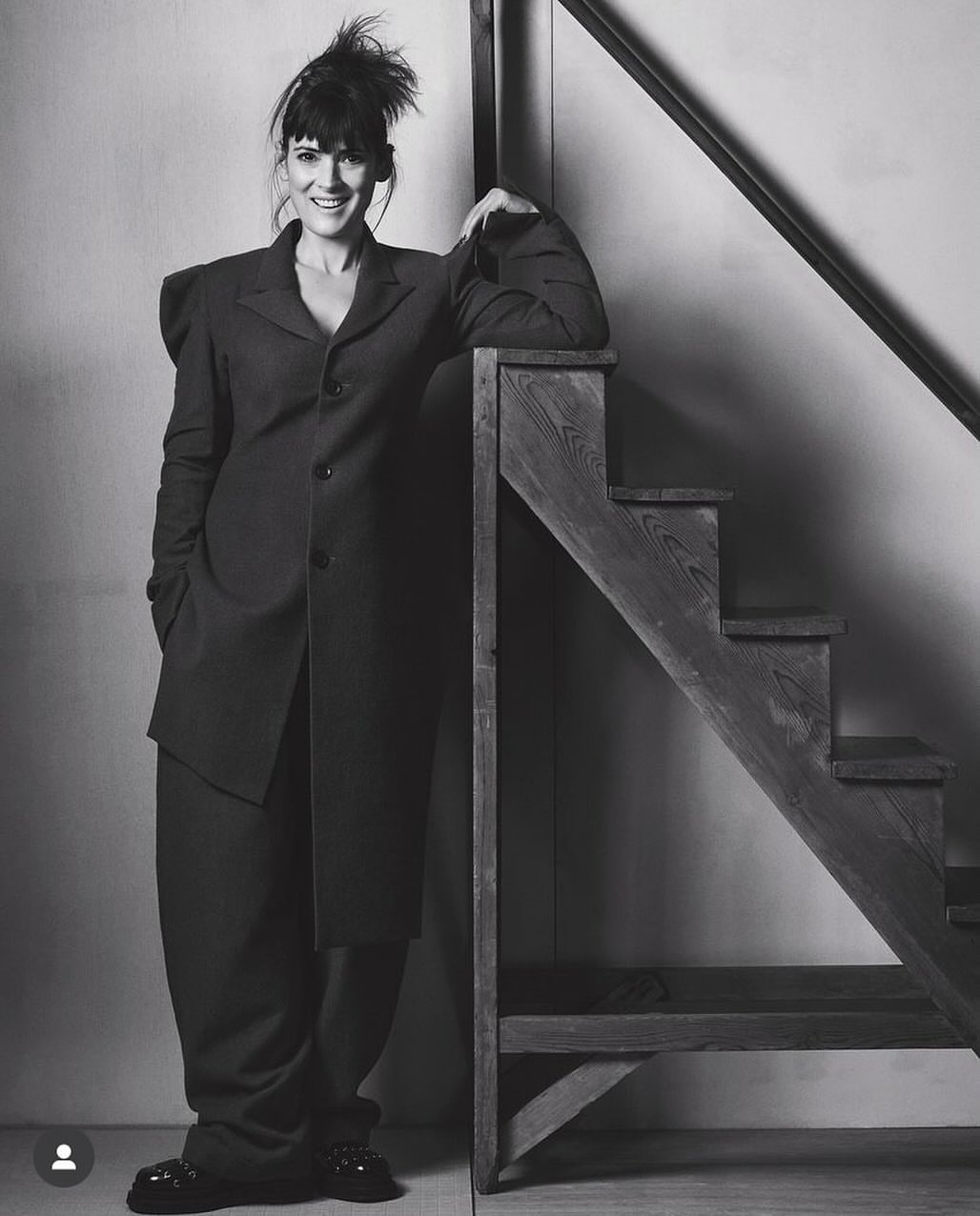 Elegant woman in a dark, oversized suit stands confidently on a wooden staircase. Monochrome studio shot.