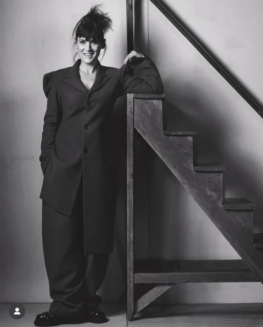 Elegant woman in a dark, oversized suit stands confidently on a wooden staircase. Monochrome studio shot.