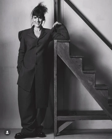 Elegant woman in a dark, oversized suit stands confidently on a wooden staircase. Monochrome studio shot.