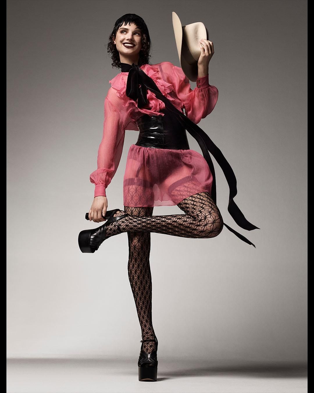 Woman in a pink dress and patterned tights strikes a pose, holding a cowboy hat, with platform shoes, against a neutral backdrop.