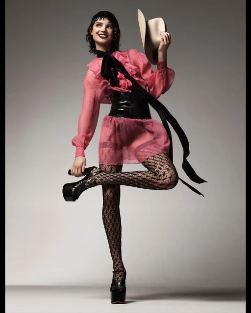 Woman in a pink dress and patterned tights strikes a pose, holding a cowboy hat, with platform shoes, against a neutral backdrop.