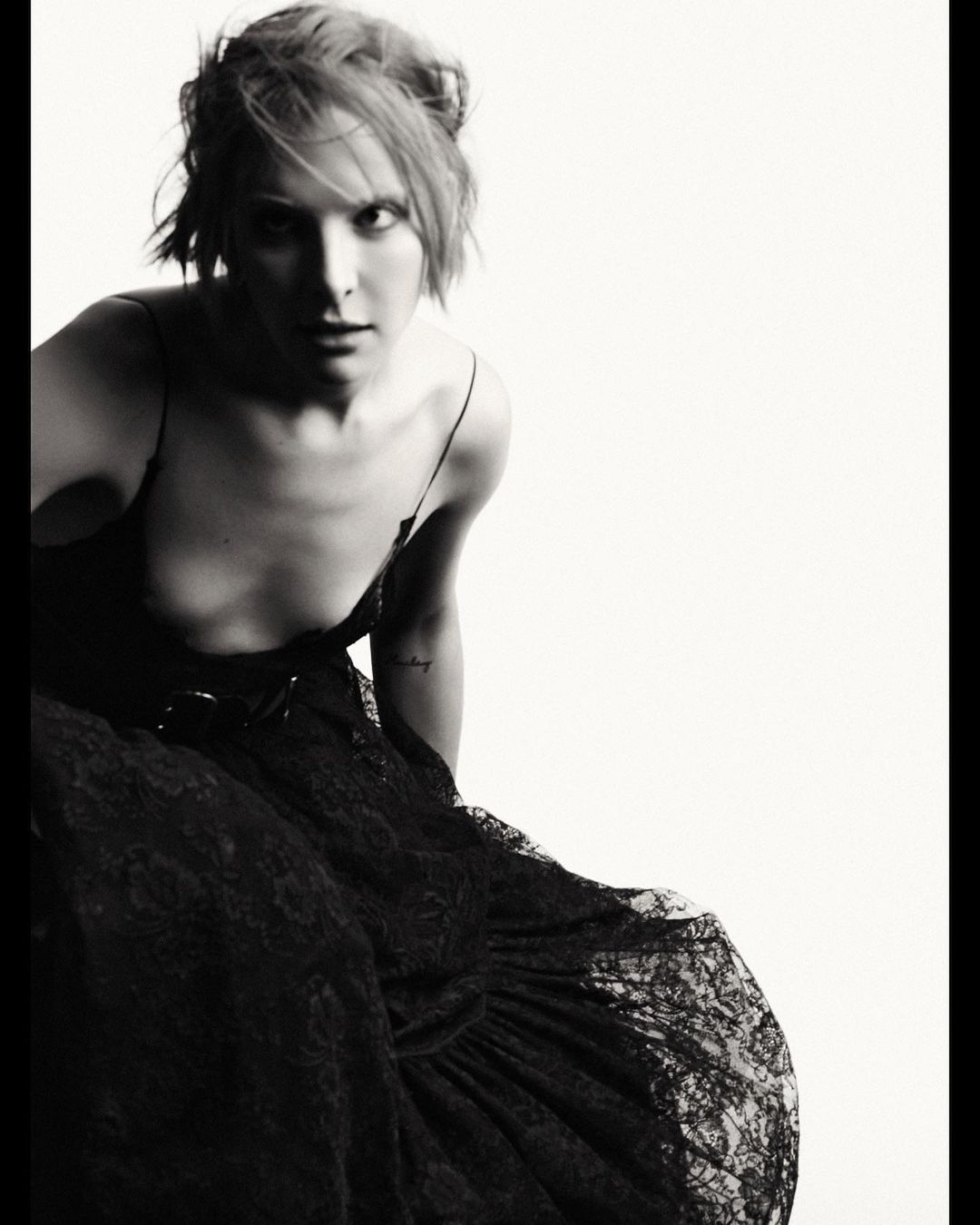 Black-and-white portrait of a woman in a black dress with a lace skirt, looking at the camera against a bright white background.