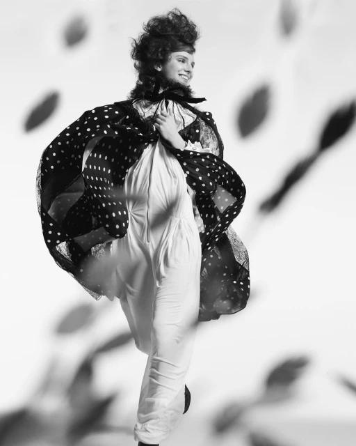 Elegant woman poses in polka dot cape and white jumpsuit against patterned backdrop in black and white fashion photography.