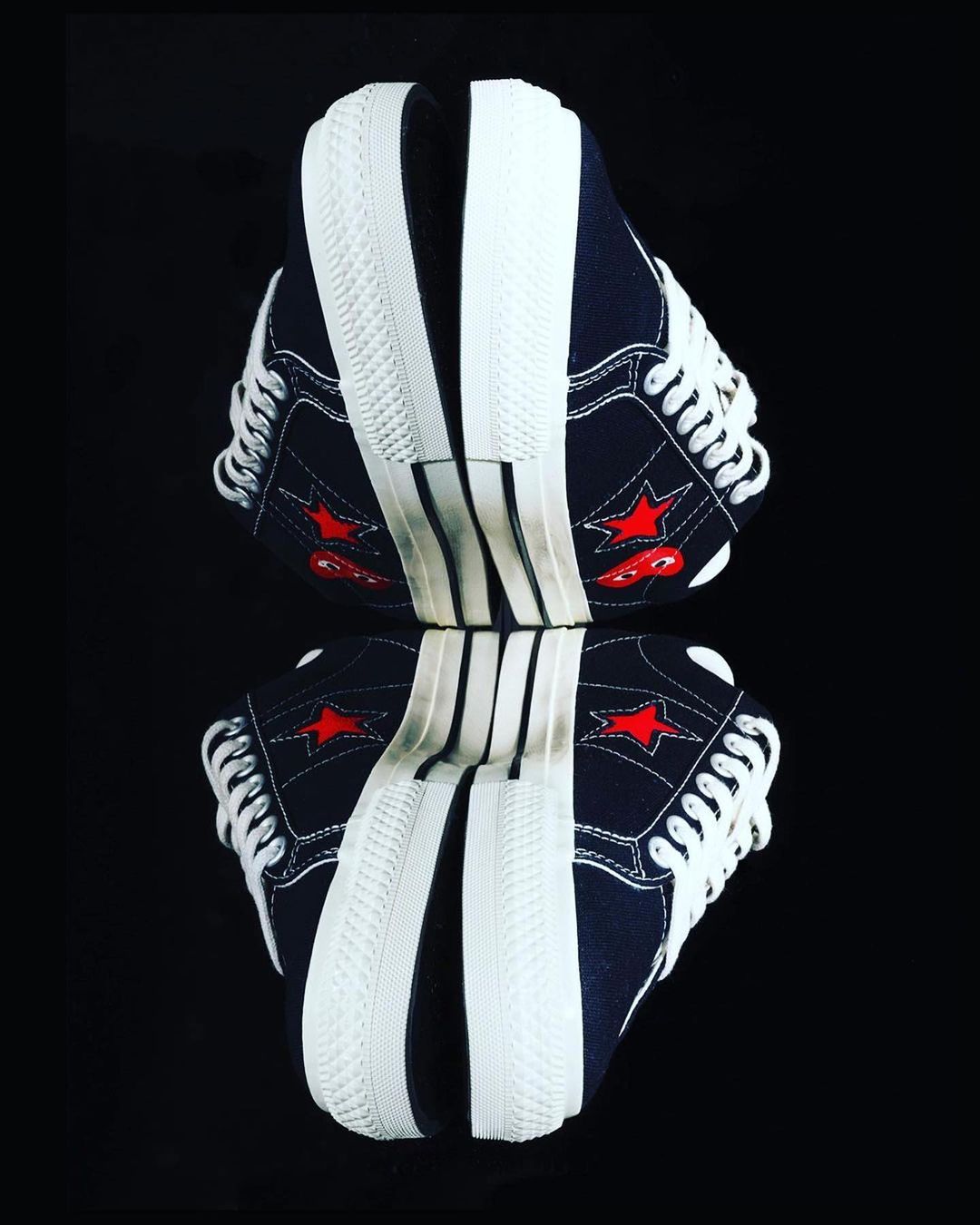 Two blue canvas sneakers with white soles and laces face each other, mirrored on a black reflective surface with a shallow depth of field.