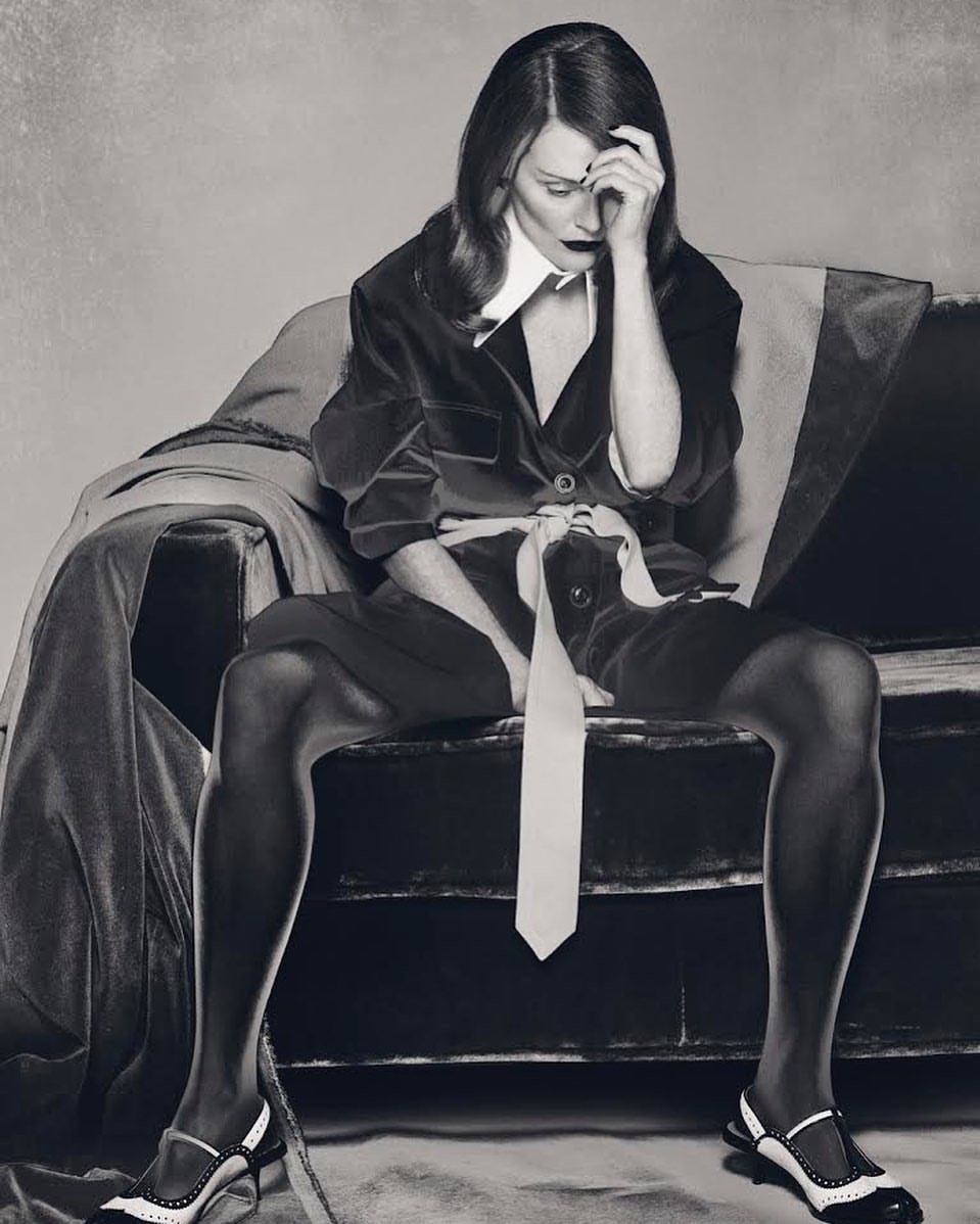 A stylish adult woman sits on a vintage sofa in a monochrome studio shot wearing black jacket, shorts, white shirt, tie, and unique heels.