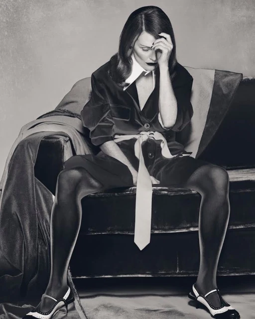 A stylish adult woman sits on a vintage sofa in a monochrome studio shot wearing black jacket, shorts, white shirt, tie, and unique heels.