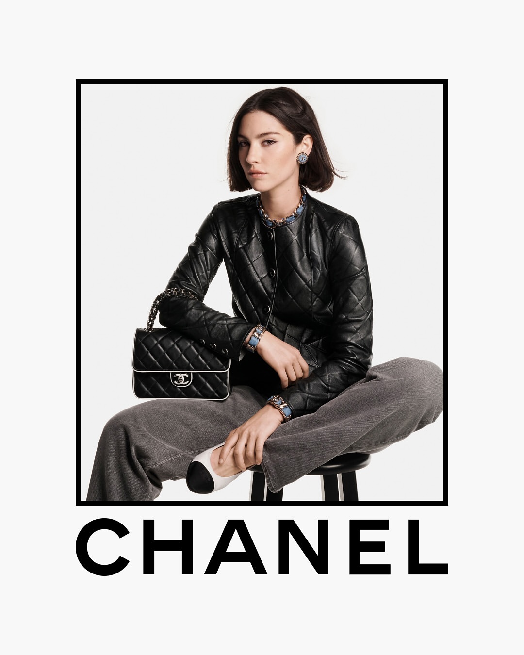 A young woman in a black Chanel jacket and grey pants sits on a stool, holding a Chanel handbag in a studio shot for a fashion advertisement.