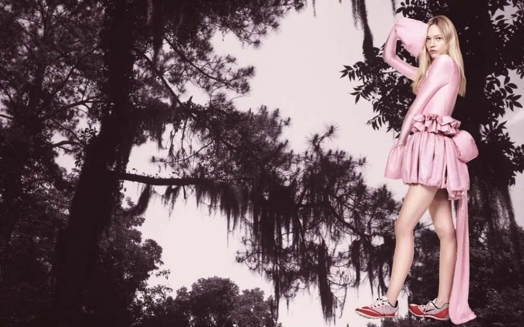 A blonde woman in a pink, frilly dress poses in front of a grayscale forest. She's wearing running shoes and holding a matching pink hat.