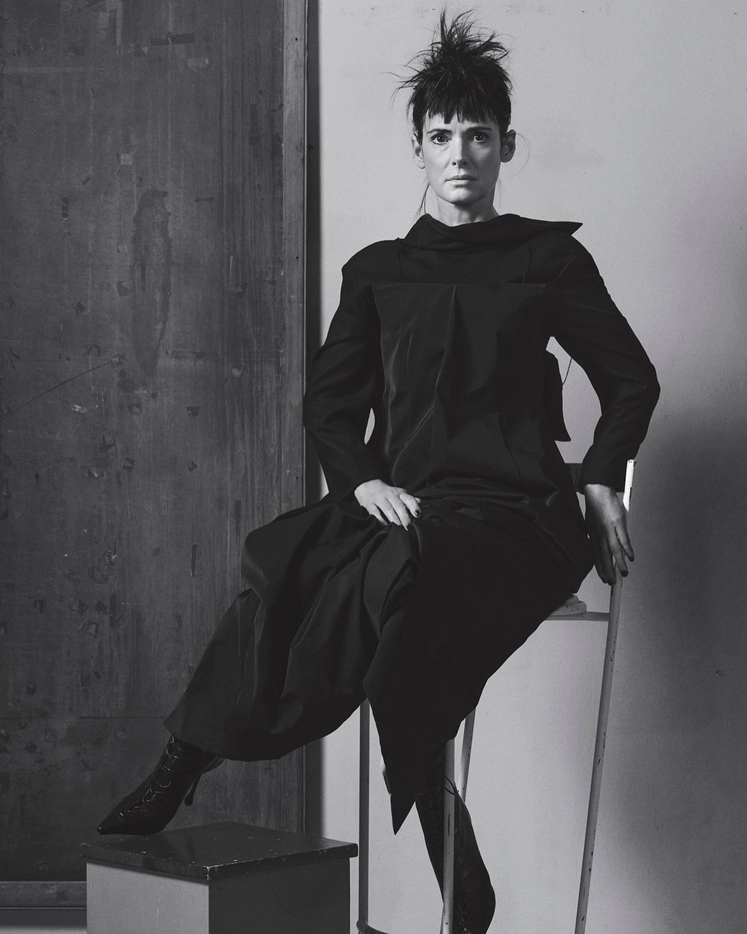Black-and-white portrait of a woman in a dark outfit seated on a chair in front of a dark panel, showcasing a blend of fashion and fine art.