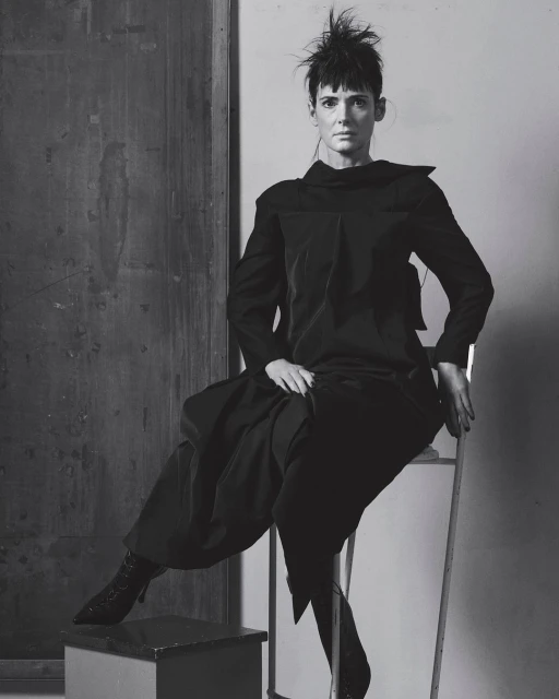 Black-and-white portrait of a woman in a dark outfit seated on a chair in front of a dark panel, showcasing a blend of fashion and fine art.