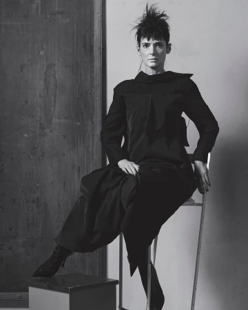 Black-and-white portrait of a woman in a dark outfit seated on a chair in front of a dark panel, showcasing a blend of fashion and fine art.