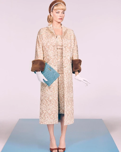 Woman wearing a patterned dress, coat, and accessories poses on a blue platform against a gradient pink and white backdrop in a studio.