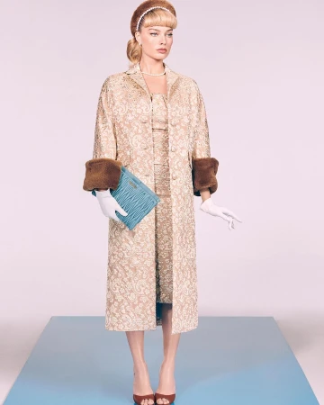 Woman wearing a patterned dress, coat, and accessories poses on a blue platform against a gradient pink and white backdrop in a studio.