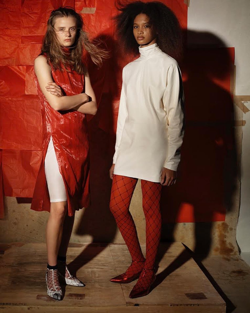 Two fashion models in a studio with a red backdrop. One wears a shiny red dress and the other an off-white dress with red tights and heels.
