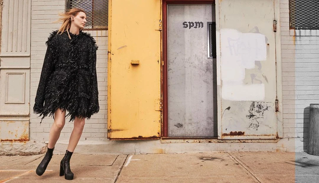 A blond woman in a textured black jacket and boots poses in front of a yellow and silver door on a city street.