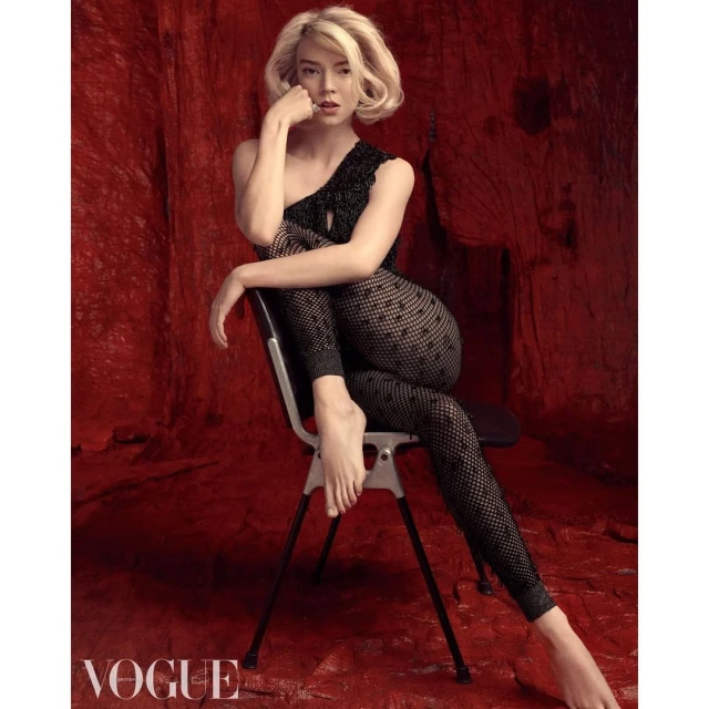 Anya Taylor-Joy is sitting on a chair in a black, polka-dot, one-shoulder jumpsuit against a red backdrop in a studio fashion shot.