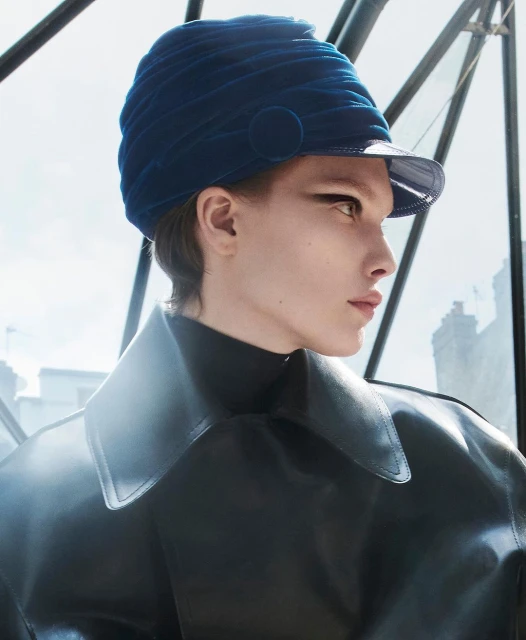 Adult female in black coat and blue hat, shot outdoors with city buildings in background, demonstrates editorial fashion photography.