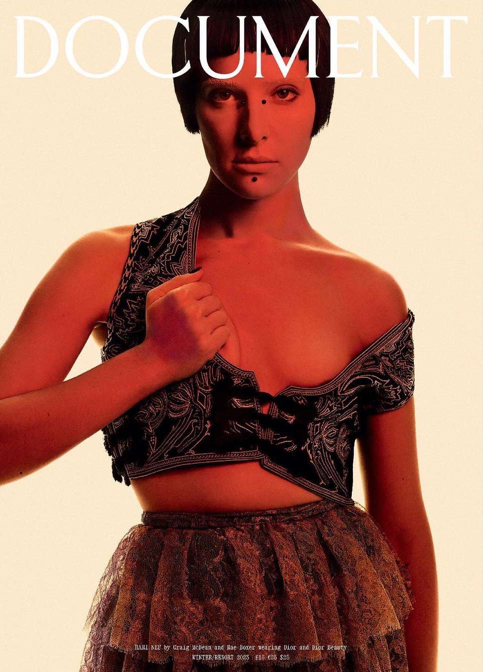A woman with a short dark bob is featured in a fashion editorial wearing a black and silver top and lace skirt against a neutral backdrop.