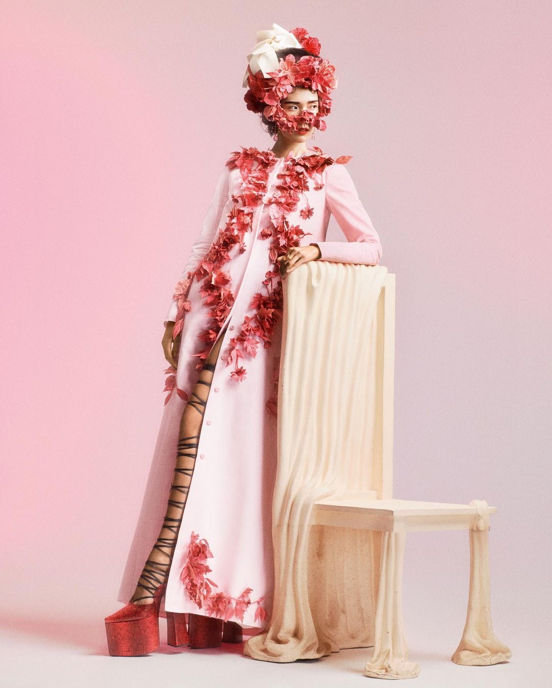 A fashion model in a light pink coat adorned with red flowers poses next to a draped chair in a pink studio setting.