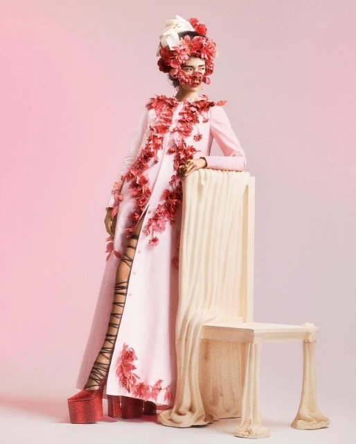 A fashion model in a light pink coat adorned with red flowers poses next to a draped chair in a pink studio setting.
