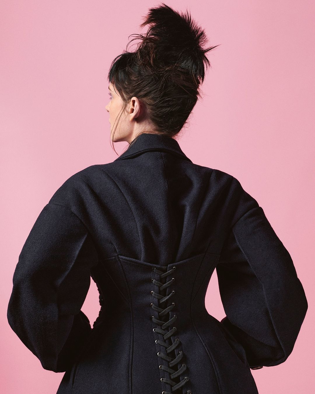Woman in a dark coat with lace-up detail posing against a pink backdrop in a studio setting, showcasing fashion and style.