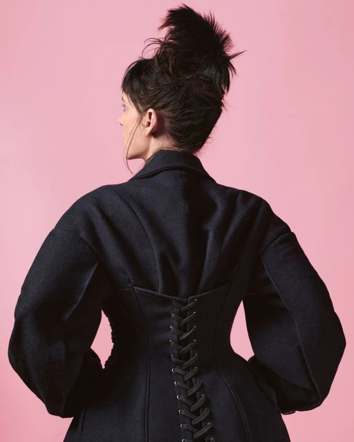 Woman in a dark coat with lace-up detail posing against a pink backdrop in a studio setting, showcasing fashion and style.