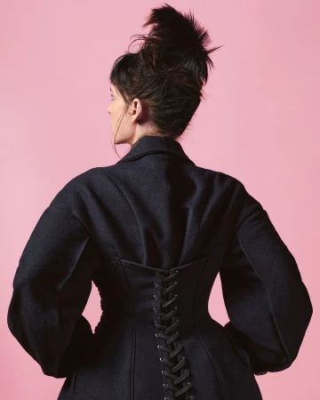 Woman in a dark coat with lace-up detail posing against a pink backdrop in a studio setting, showcasing fashion and style.