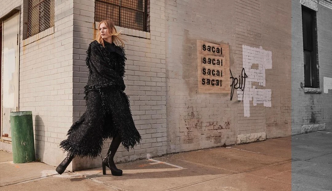A blonde woman walks in a black, fringed coat and platform boots in an urban setting by a gray brick building with graffiti.