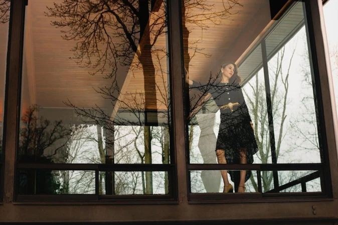 Woman in a black dress standing inside behind large windows reflecting bare tree branches