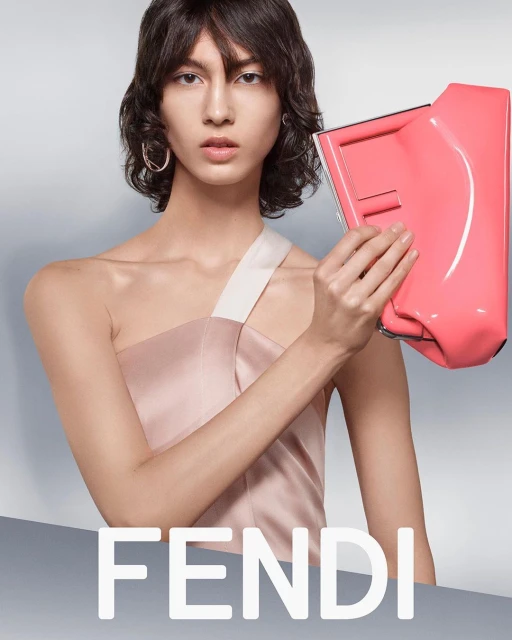 Fashion editorial shot of a woman with short dark hair holding a bright pink clutch in a studio setting. Fendi logo is visible.