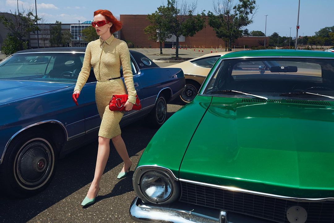 A woman in a yellow dress and red sunglasses walks past a row of vintage cars in a parking lot on a sunny day, carrying a red handbag.