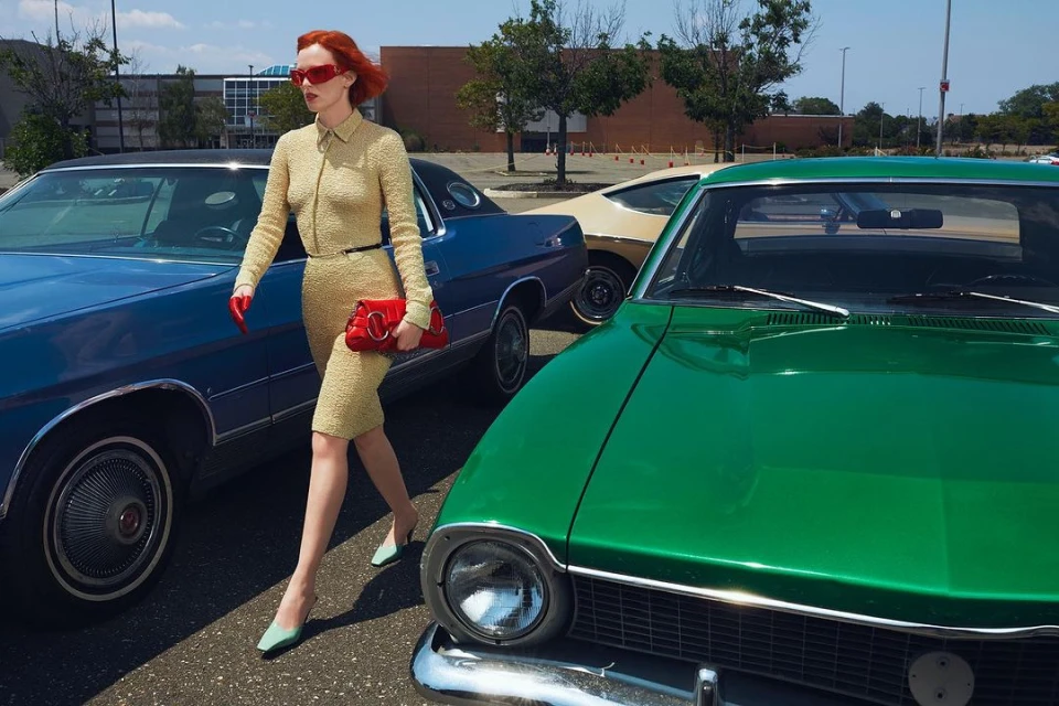 A woman in a yellow dress and red sunglasses walks past a row of vintage cars in a parking lot on a sunny day, carrying a red handbag.