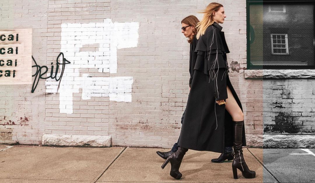 Two fashion models stride on a city sidewalk in black coats and platform boots, showcasing stylish urban attire against a graffiti wall.