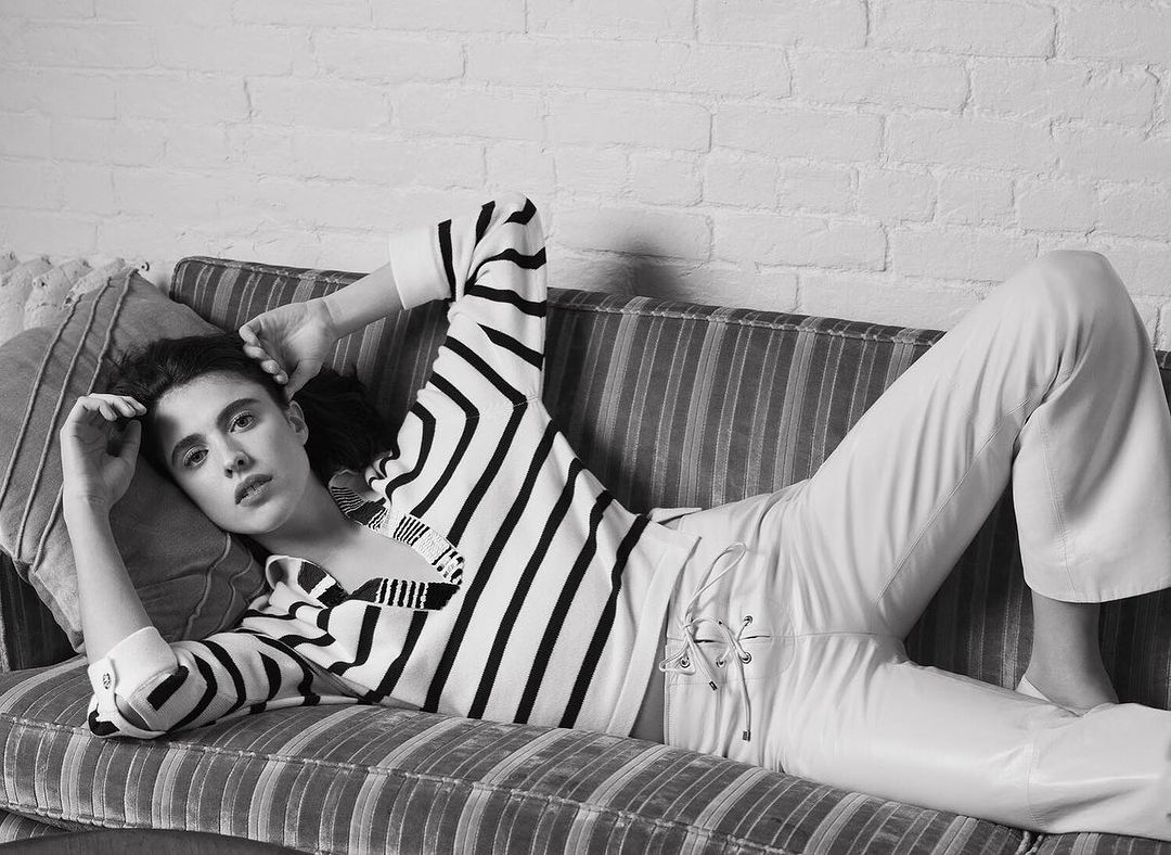 A young woman with dark hair lies on a striped couch wearing a striped shirt and white pants, looking at the camera against a brick wall.